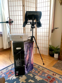 Photograph of a camera and teleprompter on a tripod stand in a living room, next to a microphone stand, in front of a transparent screen.